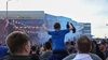 Ein junger Fan im blauen Trikot sitzt vor dem Stadion auf den Schultern eines Erwachsenen und feiert mit erhobenen Armen inmitten einer begeisterten Fangemeinde.